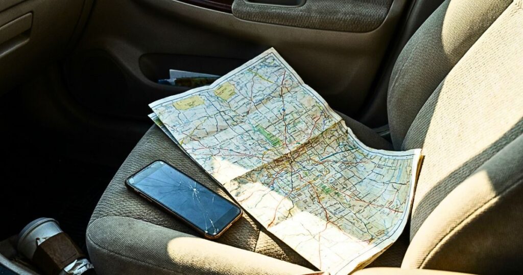 Paper road map next to a smartphone with a dark screen on a car seat