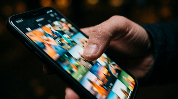 Close-up of a hand holding a smartphone with a blurred vertical video feed on the screen, thumb mid-scroll, in a dim room at night
